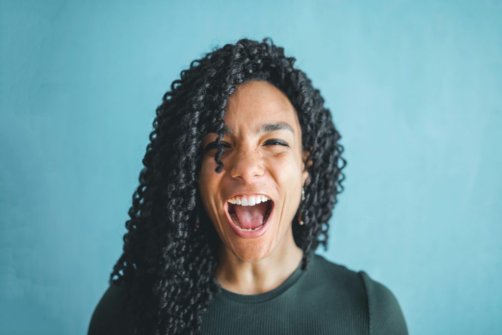 Portrait of a joyful young woman with curly hair, expressing excitement and shouting indoors.